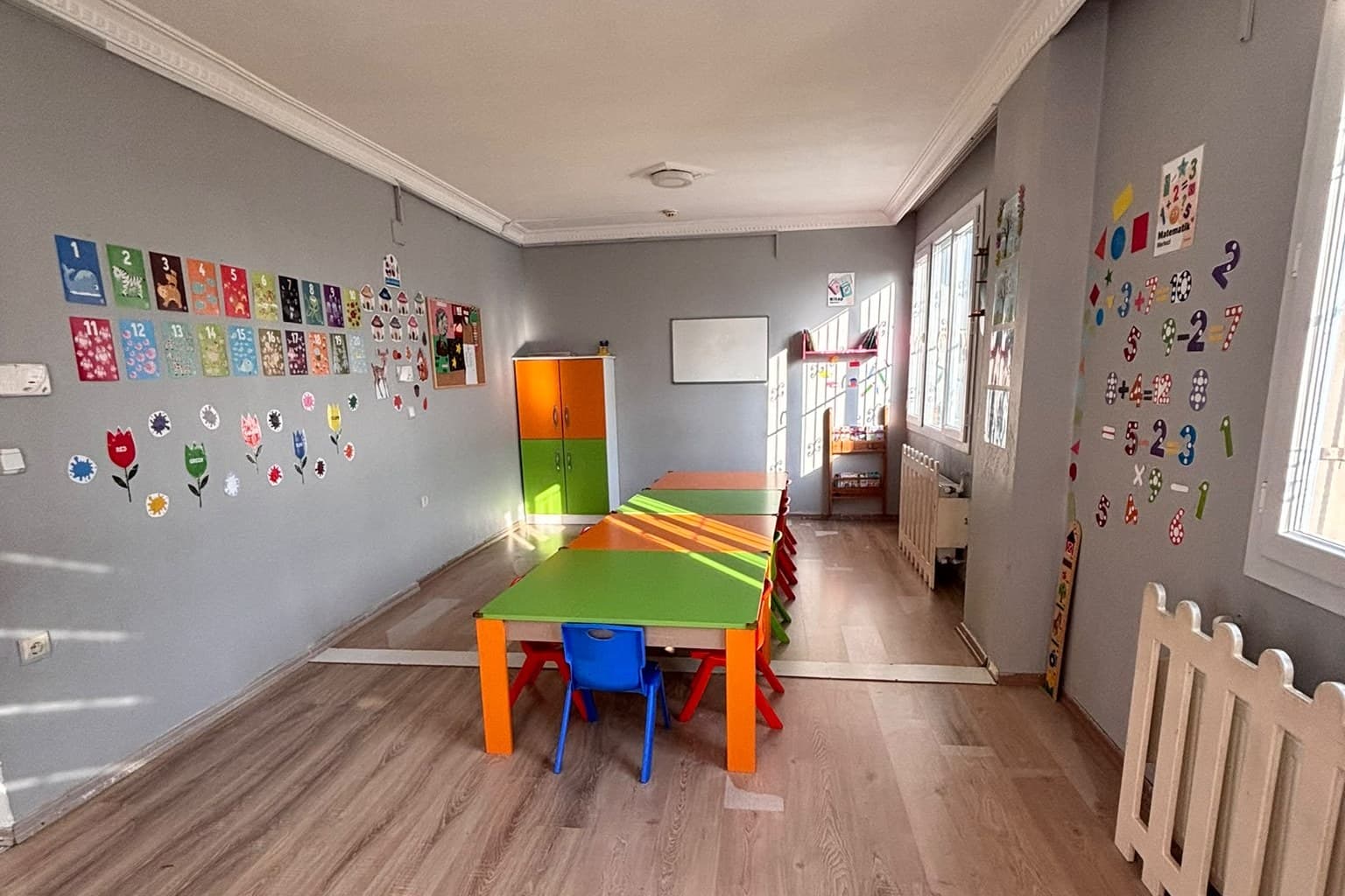 Beyaz Zambak Anaokulu sınıfı: renkli çalışma masaları ve duvarda sayı panoları